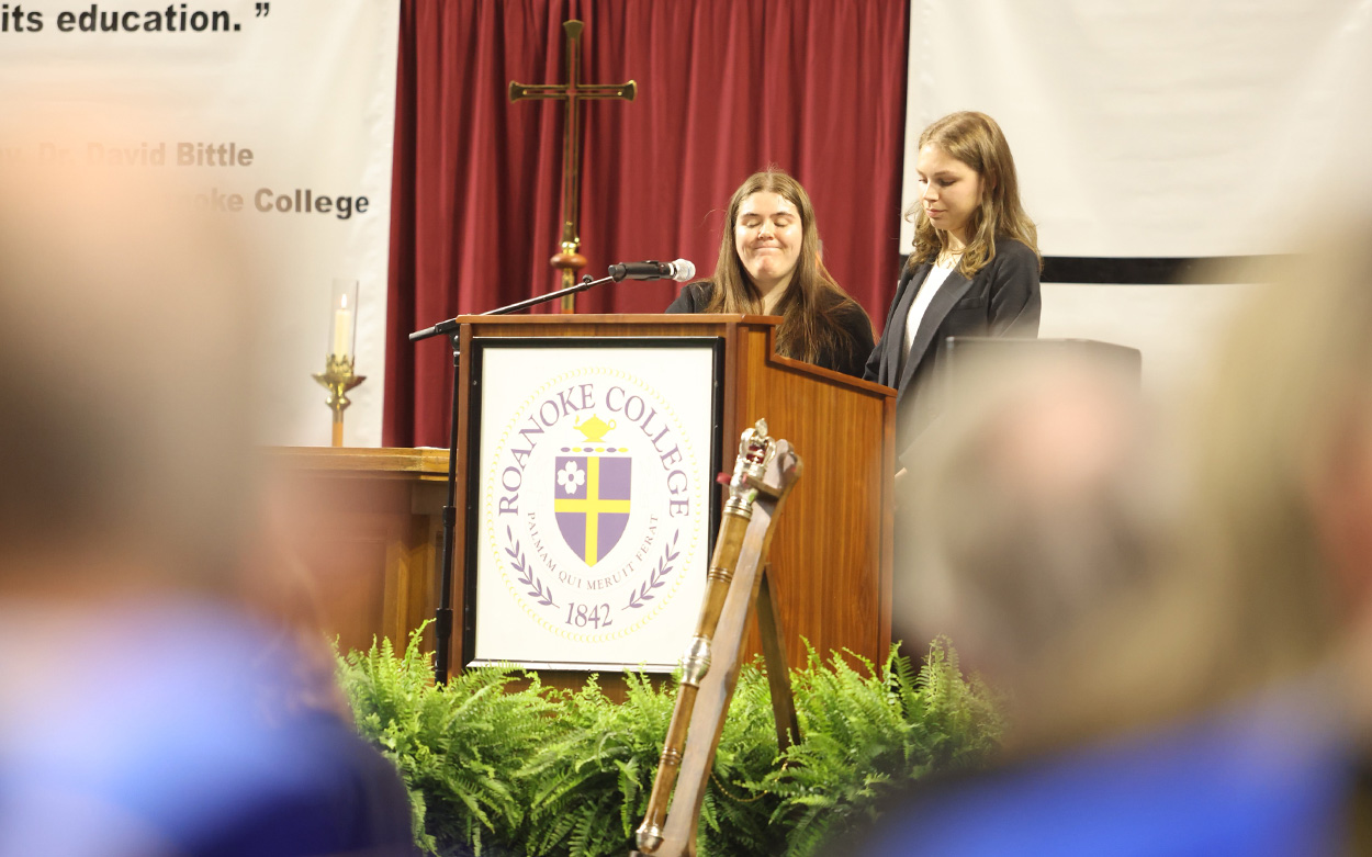 Two female students speak from a podium bearing the Roanoke College seal during an interfaith blessing