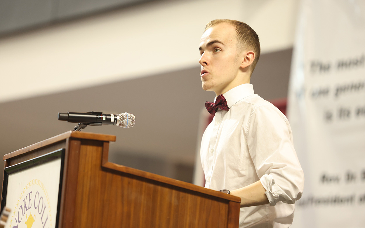 A student wearing a dress shirt and bowtie reads a poem in memory of the late Assistant Professor Mary Hill