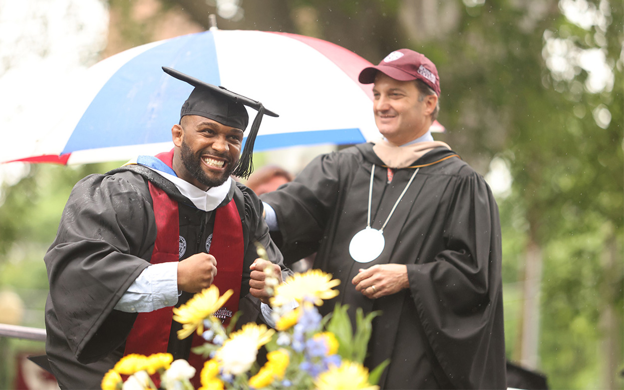 A student smiles and strikes a wrestling pose while crossing the commencement stage