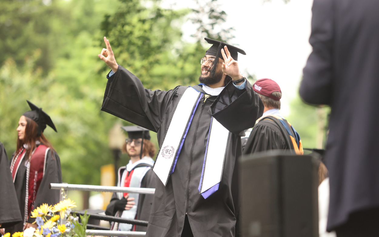 A student smiles and makes two peace signs with his hands while crossing the commencement stage to receive his diploma
