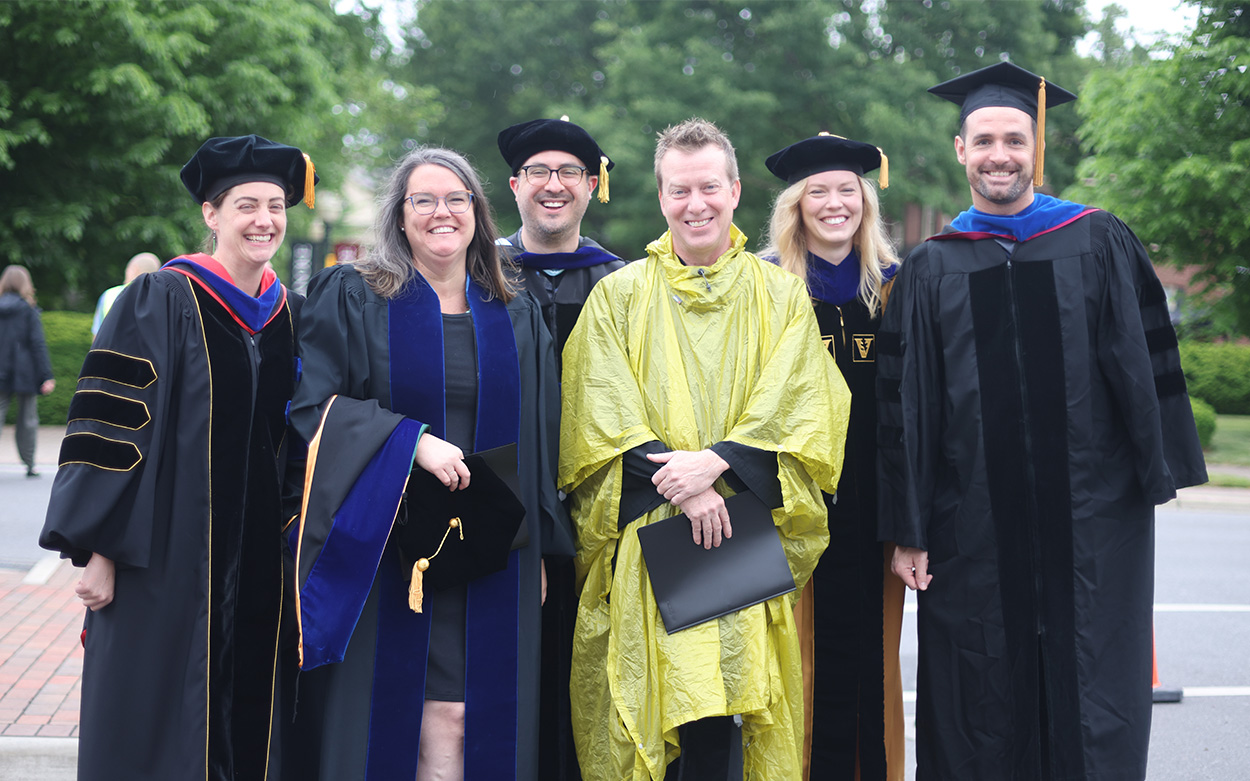 Six professors in academic robes and regalia -- and one poncho -- smile for a photo