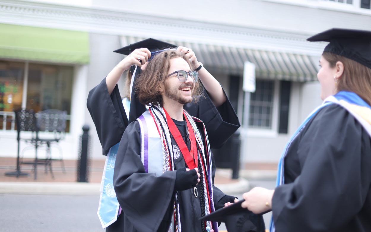 A student smiles while two friends help him don his academic regalia