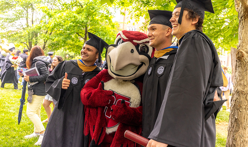 Three students smile while posing for a photo with Rooney the mascot after the commencement ceremony