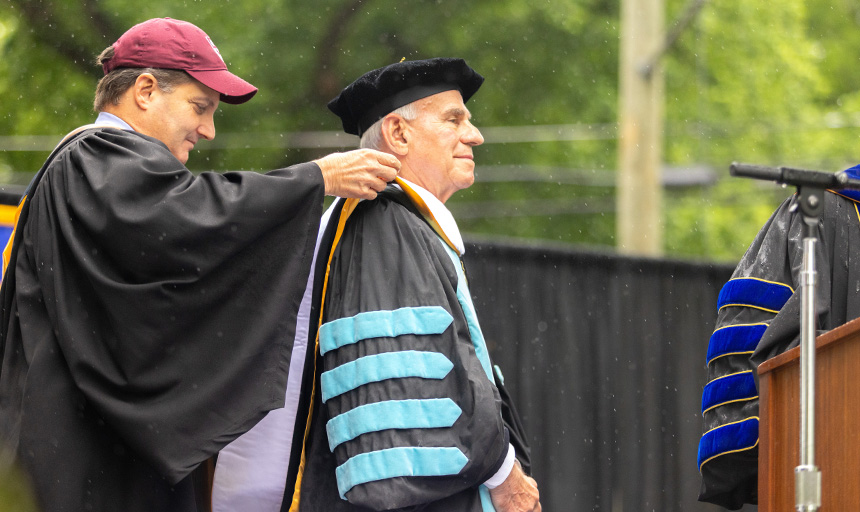 Robert Sandel, dressed in an academic robe and regalia, smiles as an honorary sash is placed around his neck