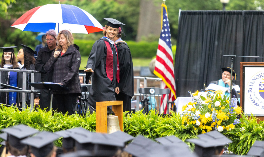A student smiles and waves while crossing the commencement stage