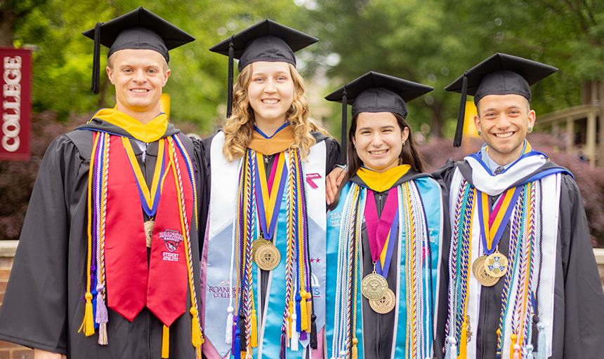 Four graduates stand, each wearing a variety of robes and honor cords