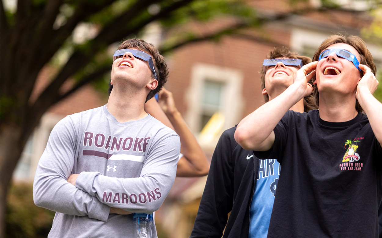 Three students wearing eclipse glasses look toward the sky