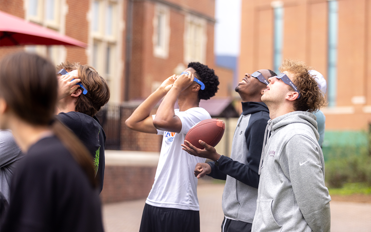 Three students wearing eclipse glasses look toward the sky while holding a football