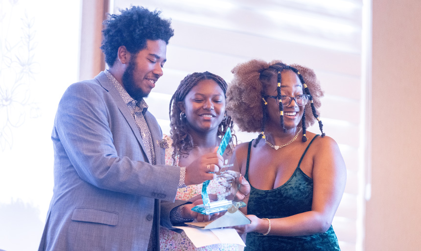 A student with emotion in her eyes smiles while being presented with an award by two other students