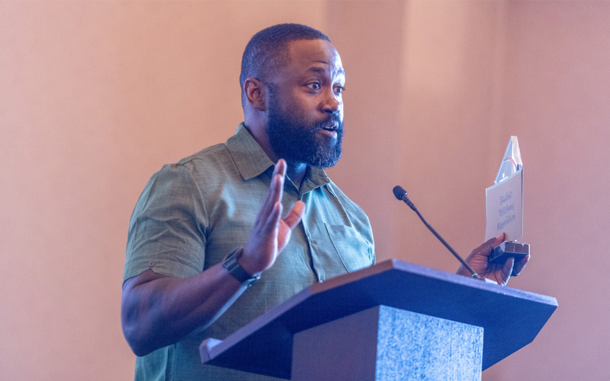 A man holds an award in one hand and gestures with the other hand while addressing the audience