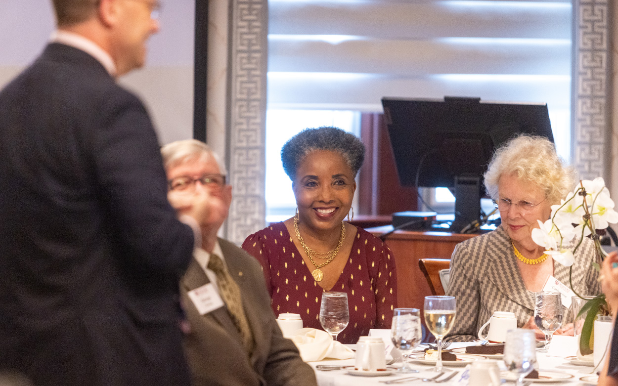 Carol Swain, seated at a reception table, smiles while a speaker addresses the room
