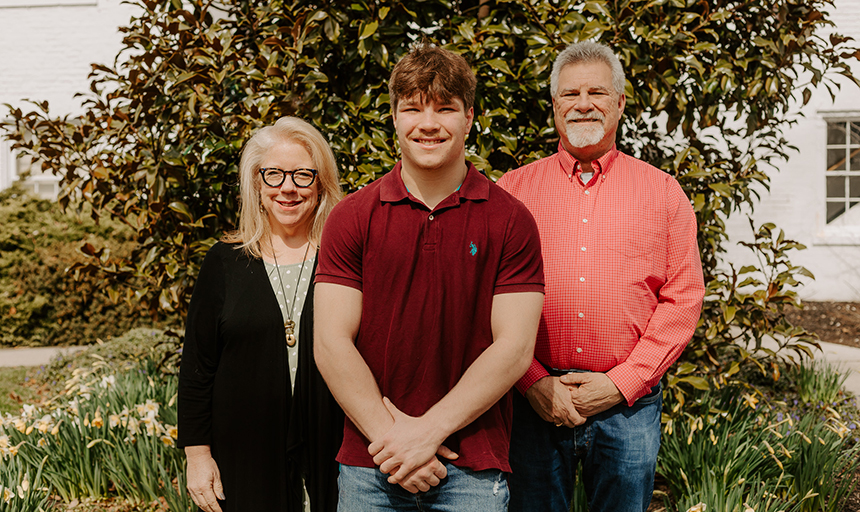 A young man in a maroon polo shirt stands in front of his parents, a blonde lady in a black sweater and a bearded man in a coral shirt, in front of a bush in what looks like their front yard.