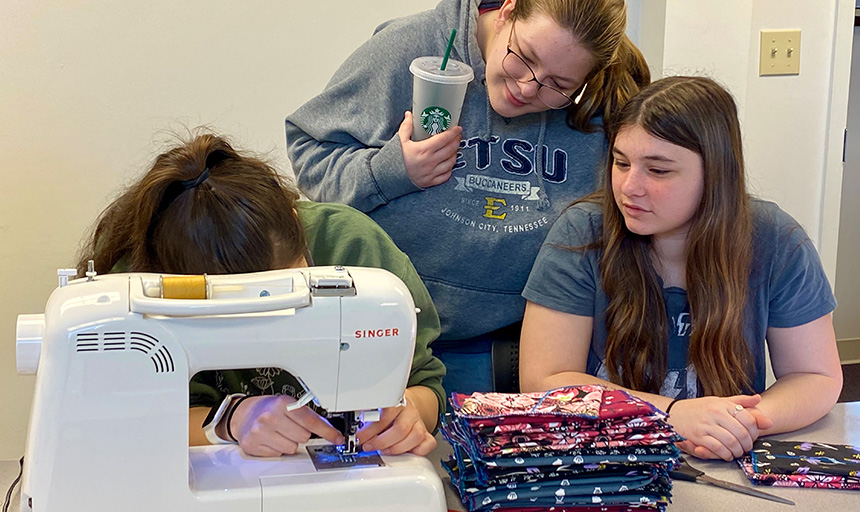 Three students gathered around a sewing machine learning to make supplies for Days for Girls kits