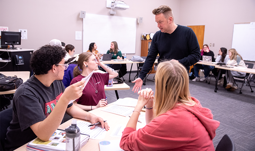 Professor stands in classroom talking to three students with others in background