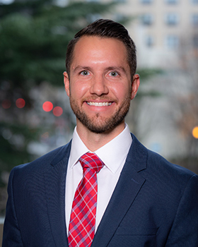 Headshot of a man in a blue suit with white shirt and red tie, dark hair and beard