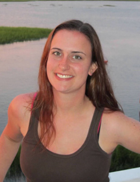 Young woman with long dark hair poses for a photo in front of a body of water at sunset or sunrise.