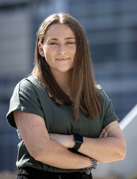 Woman with long brown hair and a dark green t-shirt poses with arms crossed in front of a blue background.