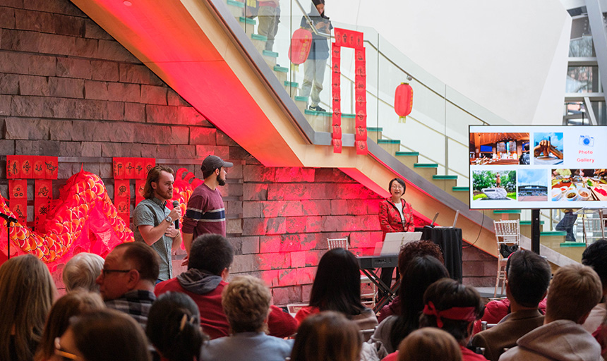 Two students and a professor address a seated audience at a museum decorated with dragon imagery and other items to celebrate Lunar New Year and the start of the year of the dragon