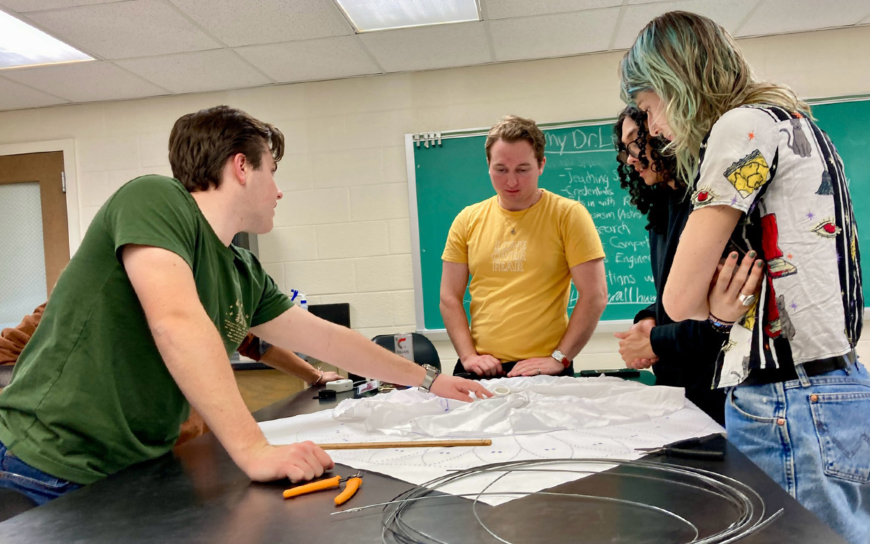 Four students stand around a waist-high work table examining a set of plans and a paper model