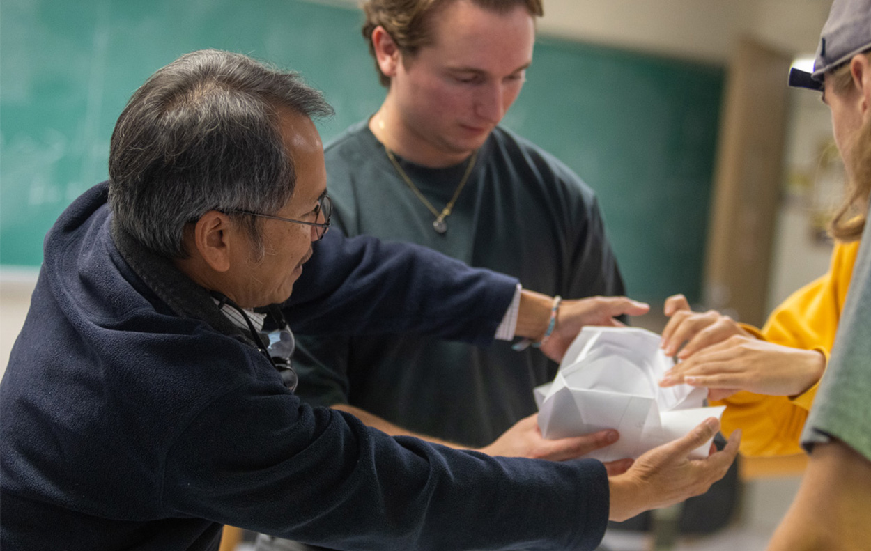 A professor helps hold a paper model aloft while students lean in to fold its edges