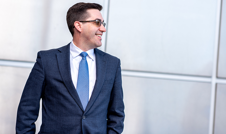 Man in a dark suit and blue tie stands in front of a silver-paneled building.