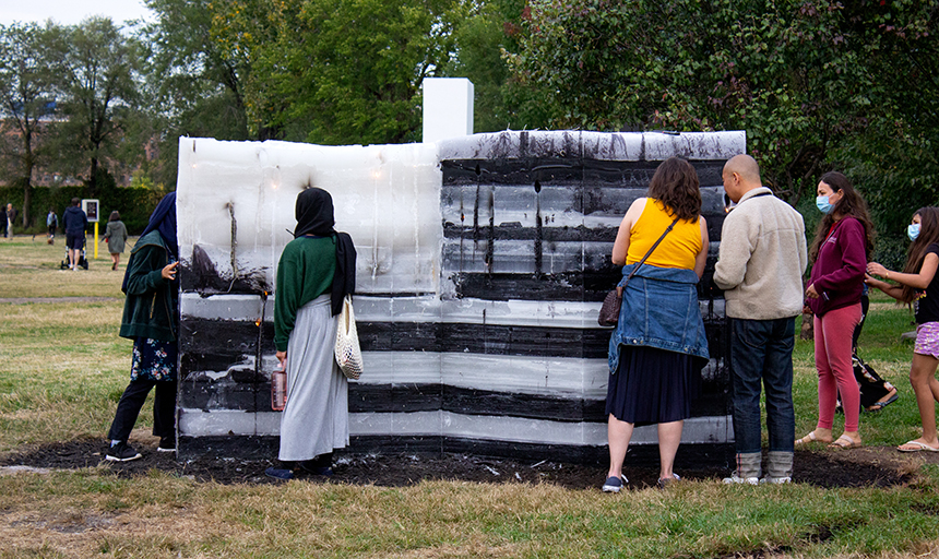 People stand around a was sculpture in a park. The sculpture looks like a black-and-white, 3D representation of the American flag. 