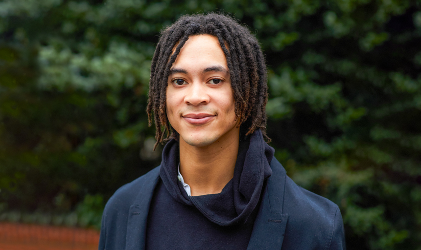 Person in a black hoodie and navy blue blazer stands in front of a wall of trees.