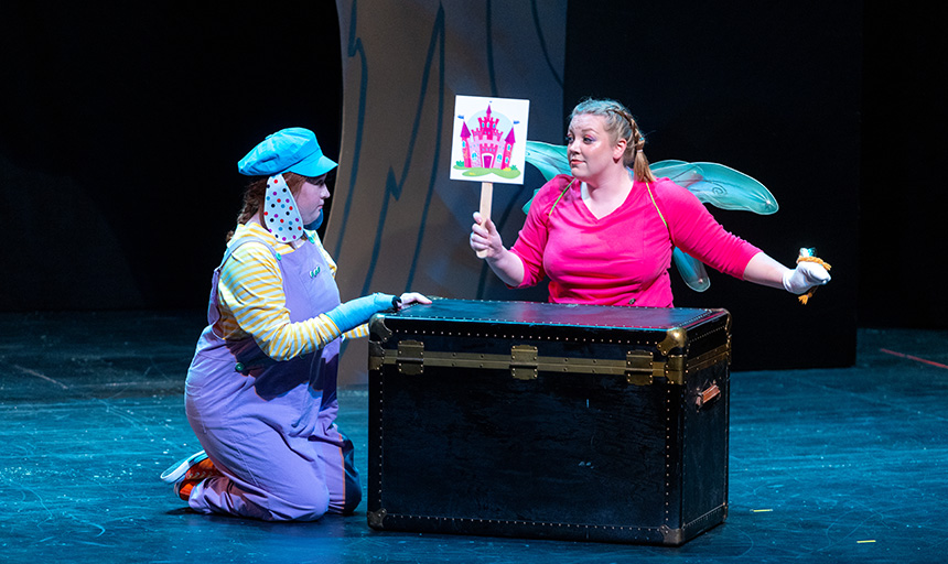 Two female actors, one dressed as a dog and one wearing fairy wings, perform on stage