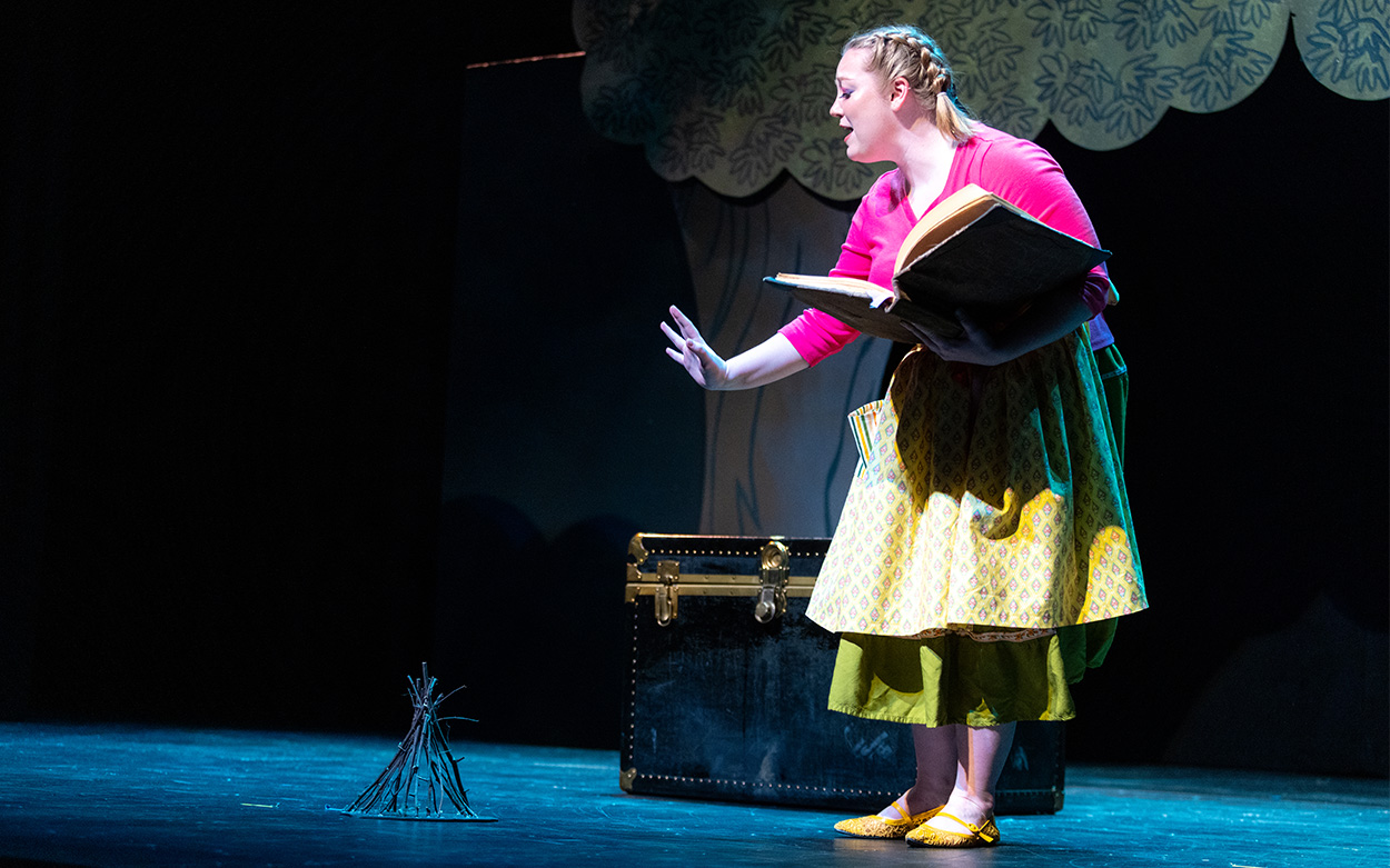 An actress on stage performs while holding a book and reaching her hand out to a stage prop of bundled twigs