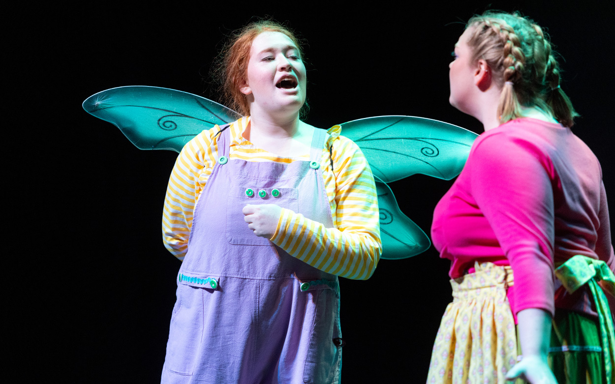 An actress wearing fairy wings speaks to another actress while performing on stage