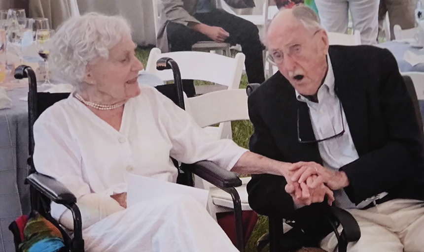 A man and woman are sitting side-by-side, and he is holding her hand and leaning toward her as he sings a song to her for their 70th anniversary.