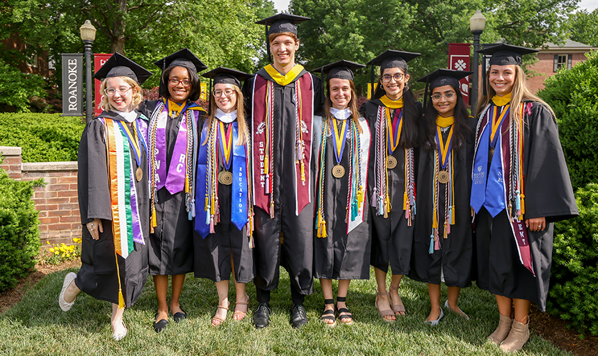 nine students in black graduation gowns with colorful tassels pose for a shot in front of a brick sign that says Roanoke College
