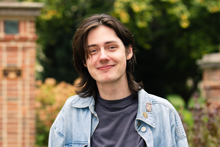 Headshot of young man with shoulder-length dark hair wearing a jean jacket