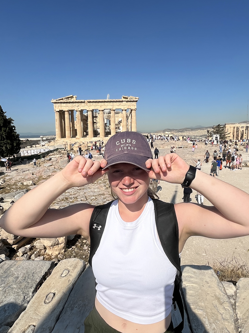 Smiling young woman in baseball cap and cropped shirt stands in front of the Parthenon in Greece
