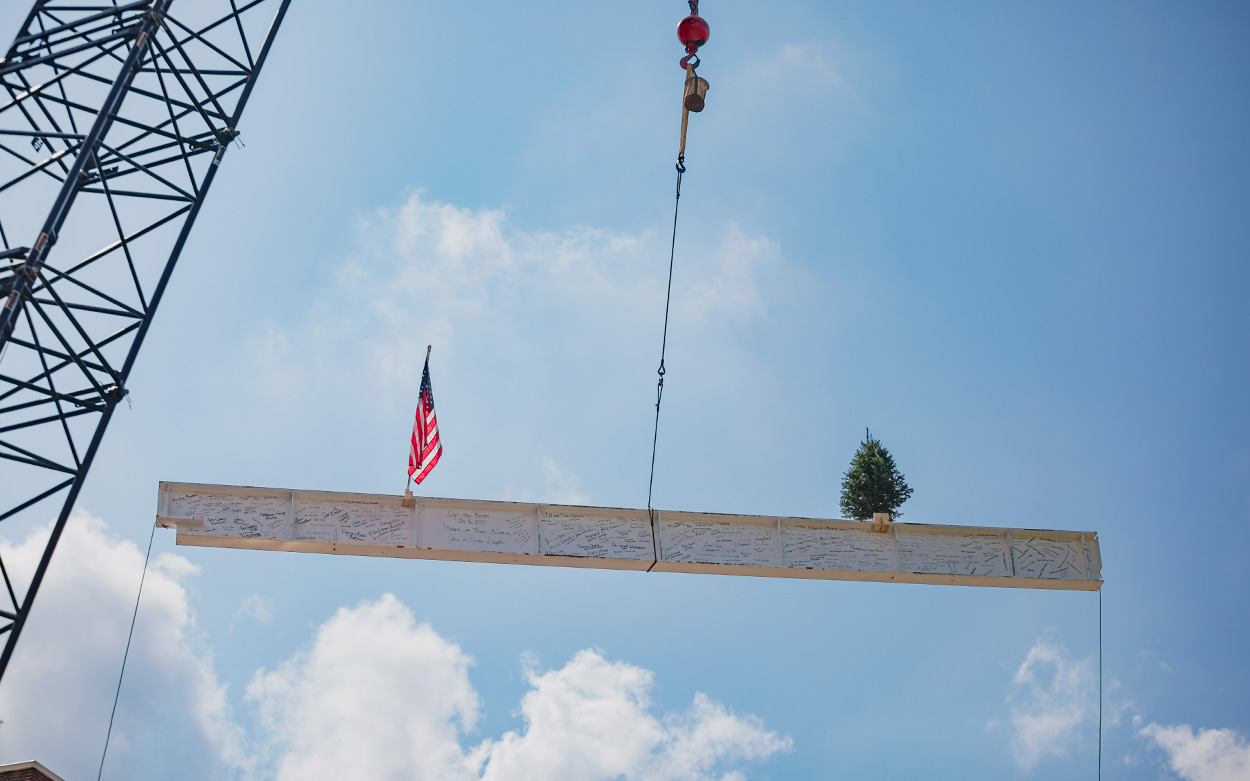 A crane hoists a steel beam that is carrying a US flag and a small evergreen tree