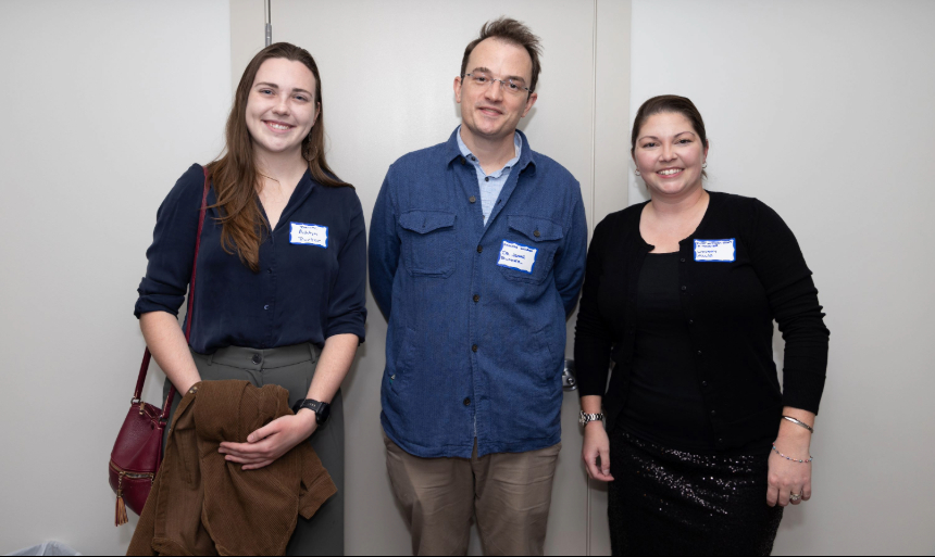 Two women and a man pose for a photo together against a white wall.