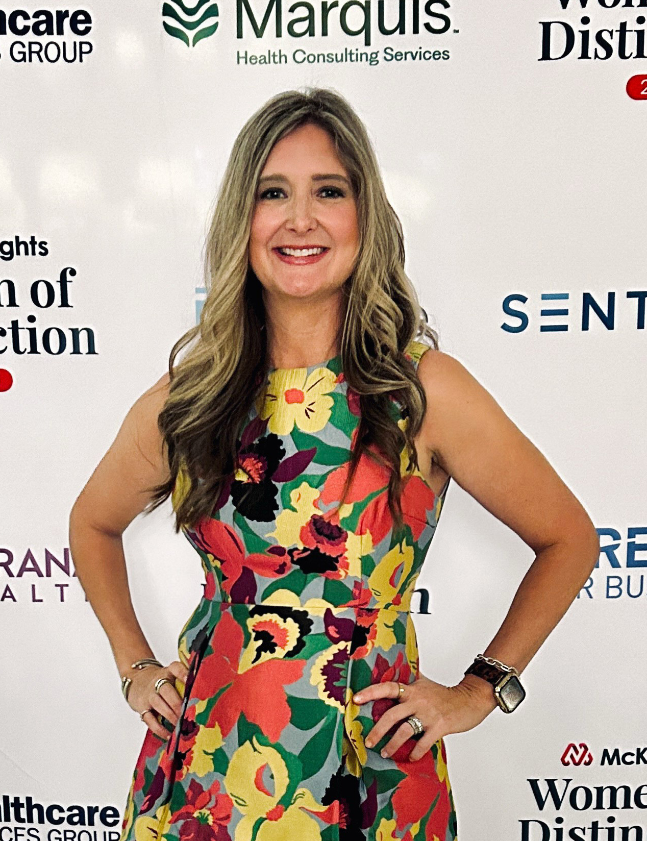 Woman in flowered dress stands with hands on hips in front of a white backdrop.