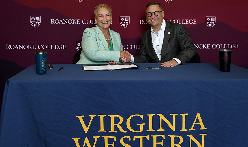 Man and a woman shake hands at a table with "Virginia Western" in yellow letters on the tablecloth, against a maroon backdrop that reads "Roanoke College."