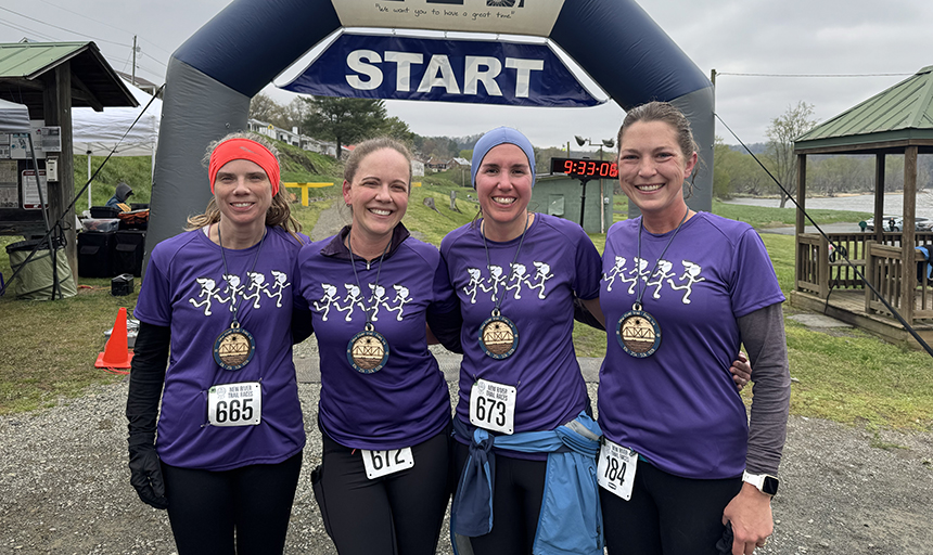 Four women in matching purple t-shirts stand arm in arm in front of the starting line for a running race.