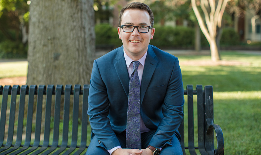 Man in spectacles and a blue suit sits on a bench with hands folded in his lap.