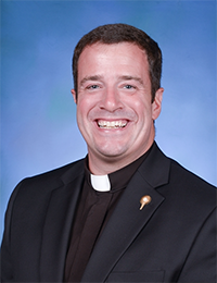 Headshot of a dark-haired man in a black shirt with white cleric's collar