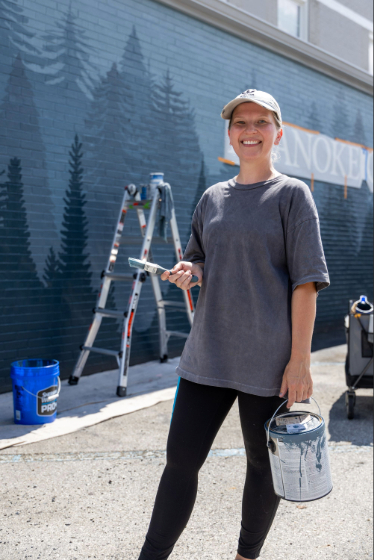 Young woman in baseball cap stands in front of a wall holding a paintbrush, and on the wall is a mural with blue mountains and trees