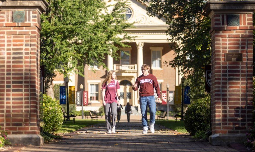 Two students walk in front of the admin building on campus. On the left is a young woman with long blond hair wearing a Roanoke College MBA shirt. The one on the right is a man with glasses and brown hair wearing a maroon sweatshirt that says "Roanoke."