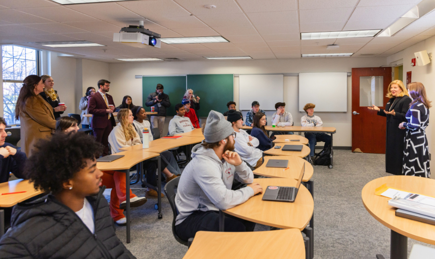 Students sit at desks inside of a classroom, all facing the front of the room where former Virginia Secretary of Education Aimee Guidera stands and speaks to them. She has blond hair and is wearing a long black dress.