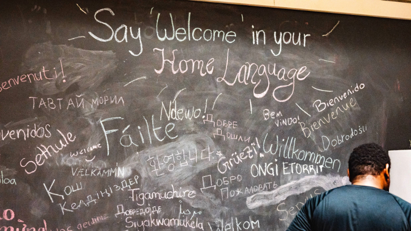 A student stands facing a chalk board that says "Say 'Welcome' in your home language." A number of responses in varying languages have been written. 