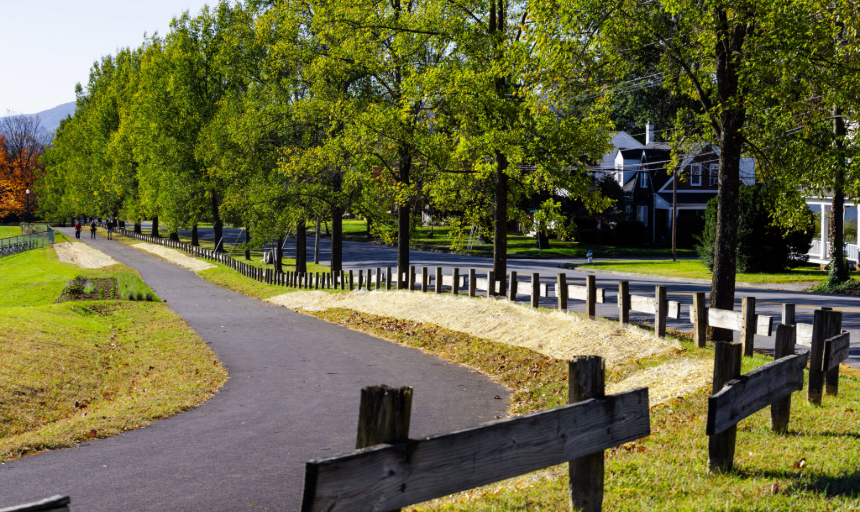 A new stretch of paved greenway on Elizabeth Campus is pictured, bordered by fences and trees. A few people are visible in the distance. 
