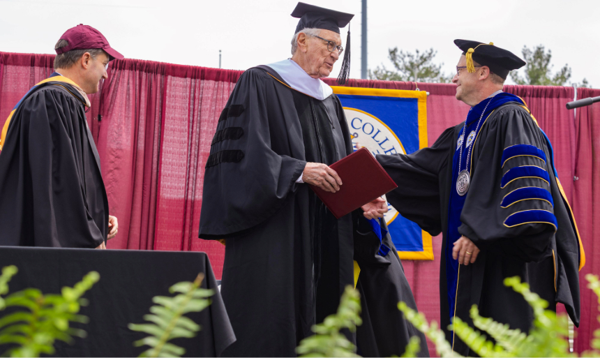 Man in black graduation-style cap and gown receives handshake from man in similar gown with blue velvet trim and cap.