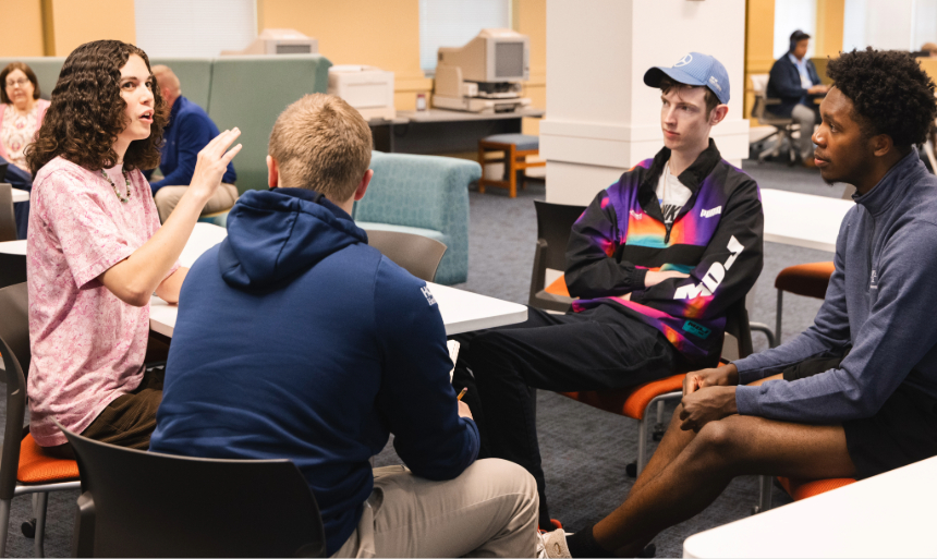 Four students sit in a circle to have a brainstorming session in Fintel Library. A student in a pink shirt with brown curly hair leads the conversation.