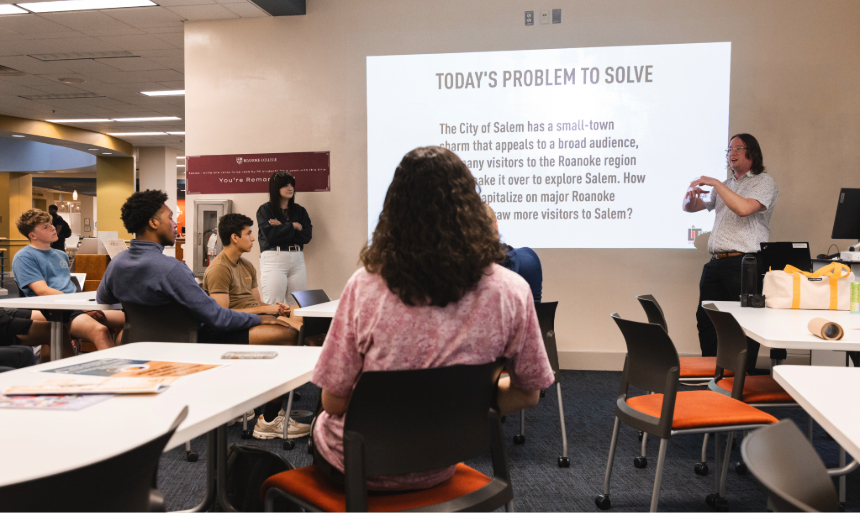 Hammerschmitt and Marsh present to a group of students in the Fintel Library. A slide is projected onto the wall behind Hammerschmitt that says "Today's problem to solve" and lists the problem Marsh is presenting from the City of Salem. 