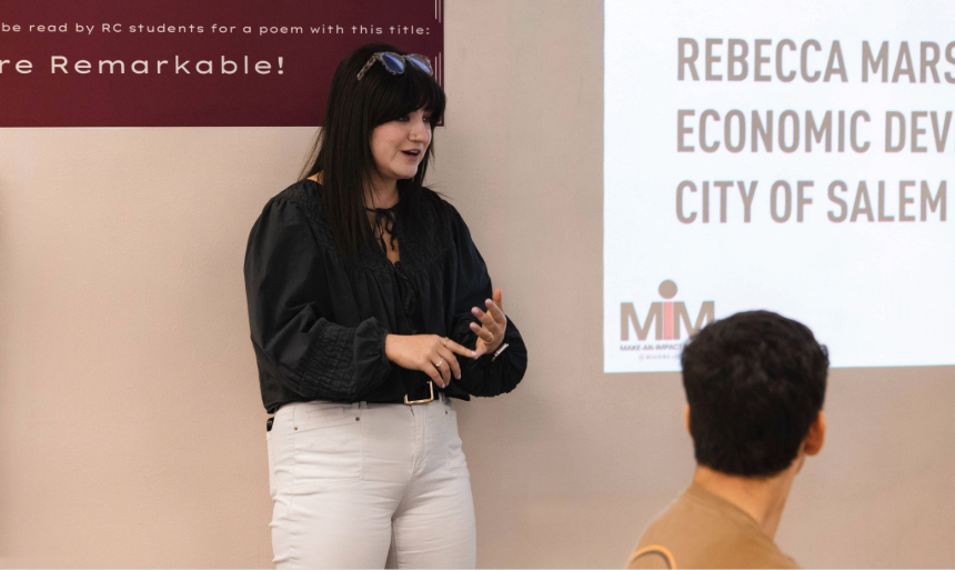 A woman with long black hair gives a presentation during Make-an-Impact Monday in the Fintel Library. She is wearing glasses on top of her head, a black long-sleeved blouse and white pants. 